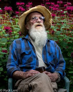 Portrait of seated older man with straw hat, glasses, white beard, and blue work shirt; looking slightly up and right; background of dark brown wood and a row of bright red bee balm flowers at top.